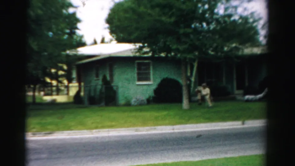 1957 home-movie frame from Normal, Illinois, showing a green single-story house, lawn, trees, and adults near the front entrance.