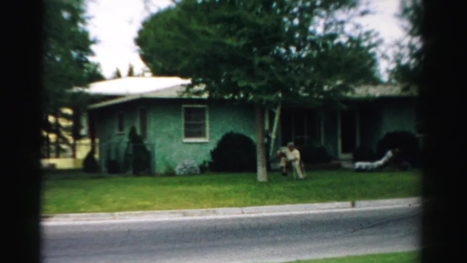 A 1957 Normal, Illinois residential exterior shows adults near a chair on the lawn before a green house with white trim.