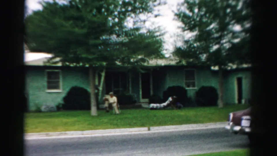 Adults are visible on the lawn of a green Normal, Illinois house while a maroon mid-century car appears at the right edge.