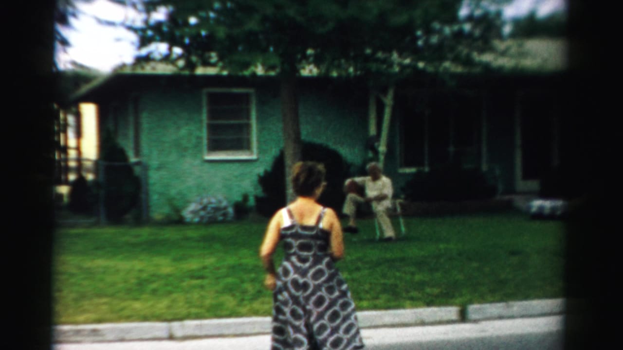 Woman in patterned dress stands near a curb facing a seated man on a lawn.