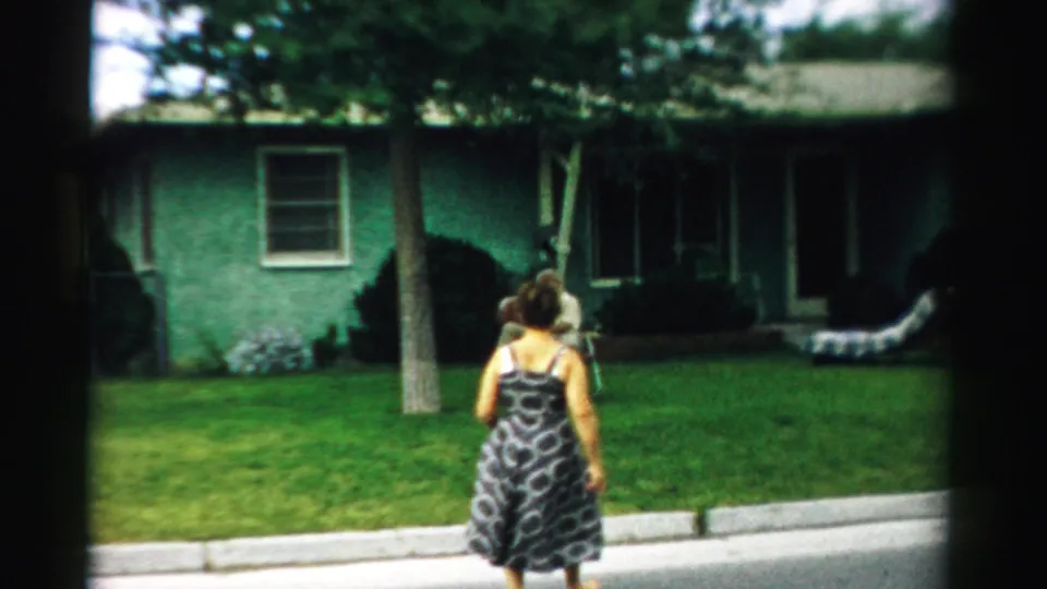 The woman continues across the lawn toward the seated man, with a green house and tree behind them in the 1957 Illinois scene.