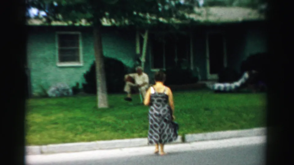 A woman in a patterned dress moves closer to a seated man on a residential lawn in Normal, Illinois, 1957.