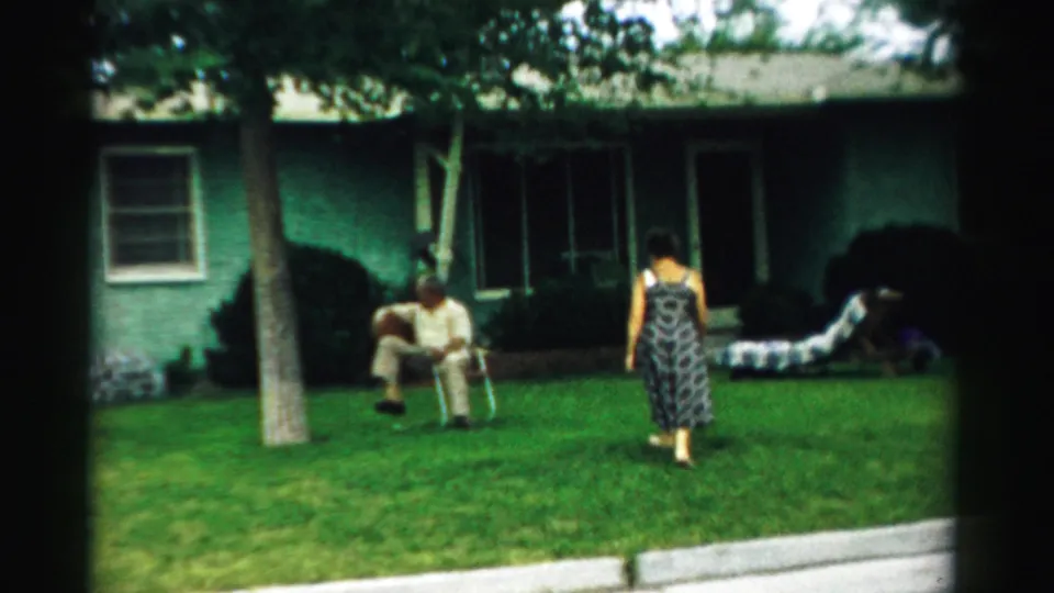 The woman crosses the front yard toward the seated man, with a lounge chair and green house visible in the background.