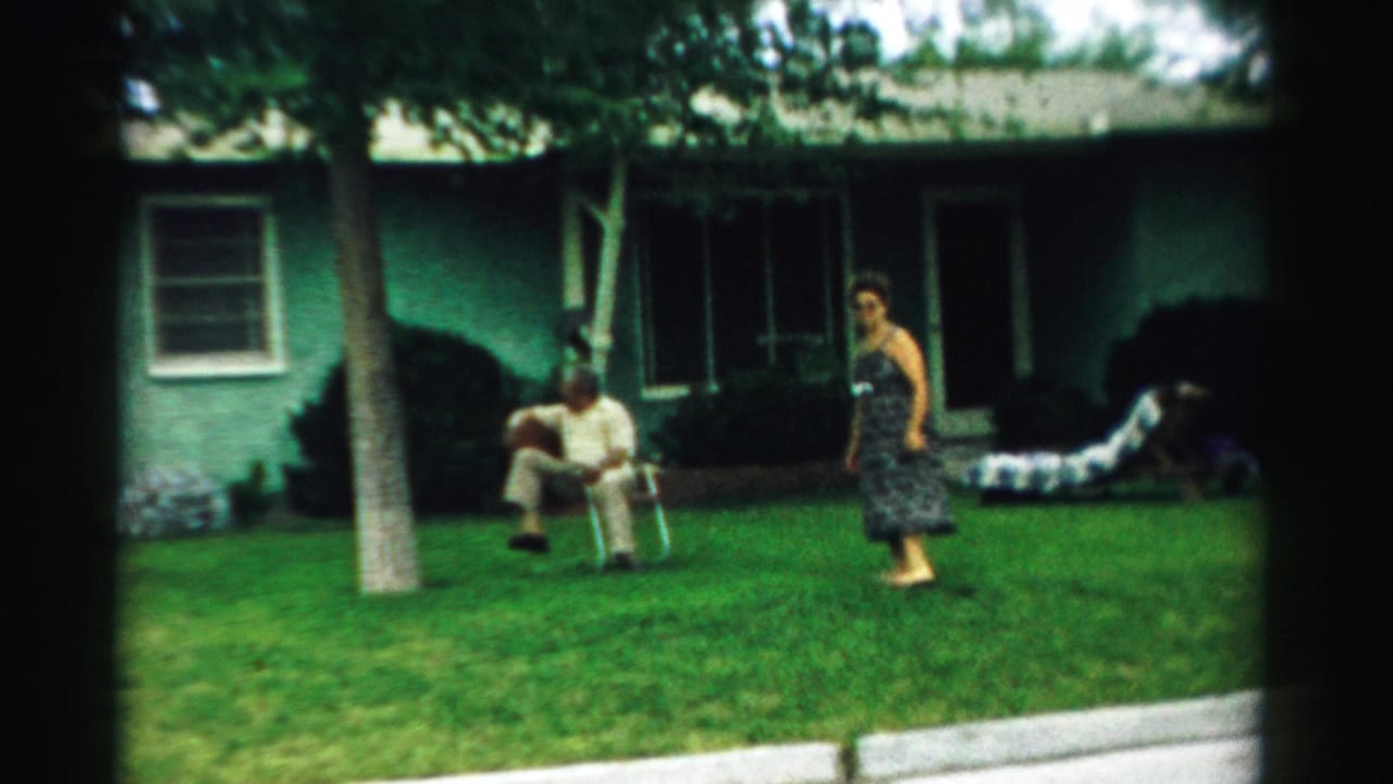 In supplied 1957 Normal, Illinois home-movie footage, an older man sits in a folding chair on a lawn as a woman in a patterned dress stands near a green stucco house.