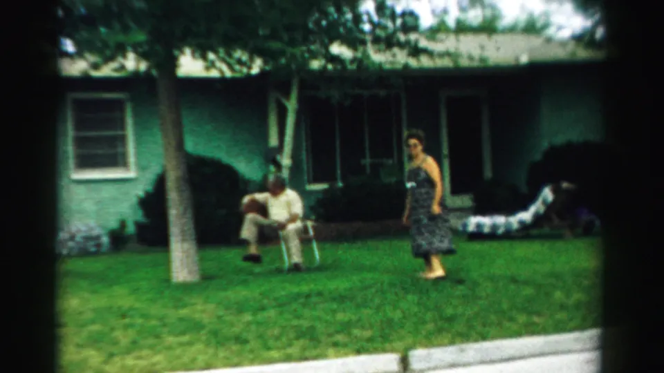 In supplied 1957 Normal, Illinois home-movie footage, an older man sits in a folding chair on a lawn as a woman in a patterned dress stands near a green stucco house.