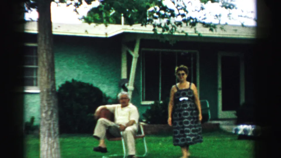A closer 8mm frame shows the older man in a light outfit seated outside as the woman in a patterned dress moves across the sunlit lawn in front of the house.