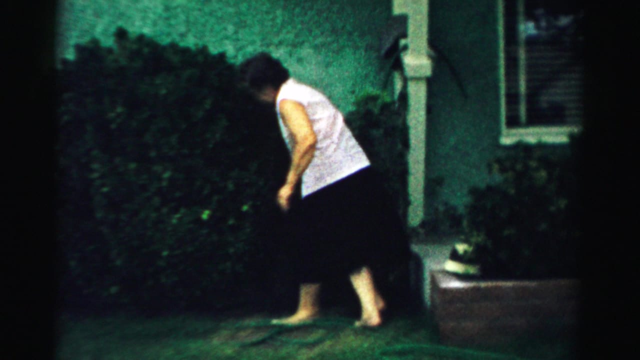 In a 1957 Normal, Illinois home-movie frame, a woman bends near shrubs beside a green stucco house exterior.