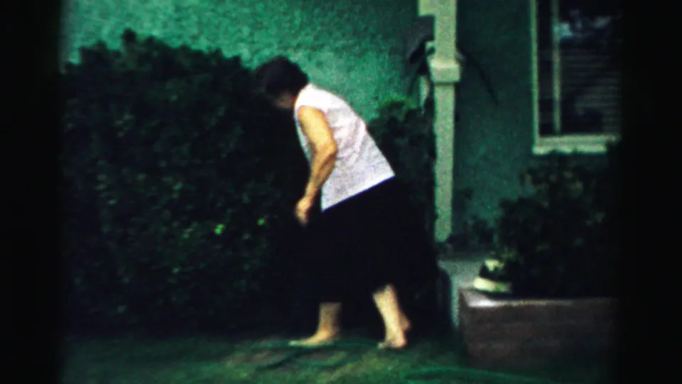 In a 1957 Normal, Illinois home-movie frame, a woman bends near shrubs beside a green stucco house exterior.