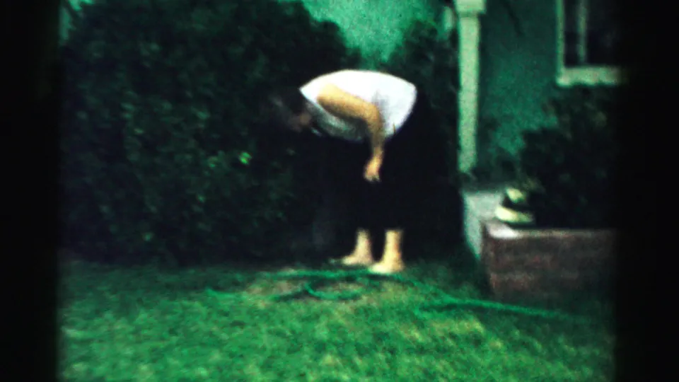 The woman bends forward on the lawn near bushes and a green house exterior in this 1957 home-movie frame.