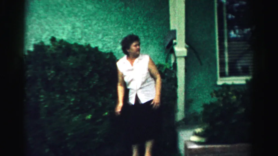 A woman stands beside shrubs, a porch post and a window in a Normal, Illinois backyard home-movie scene from 1957.