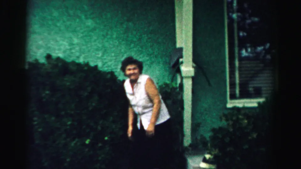 The woman appears to look toward the camera while standing by shrubs and a porch post in the 1957 backyard footage.