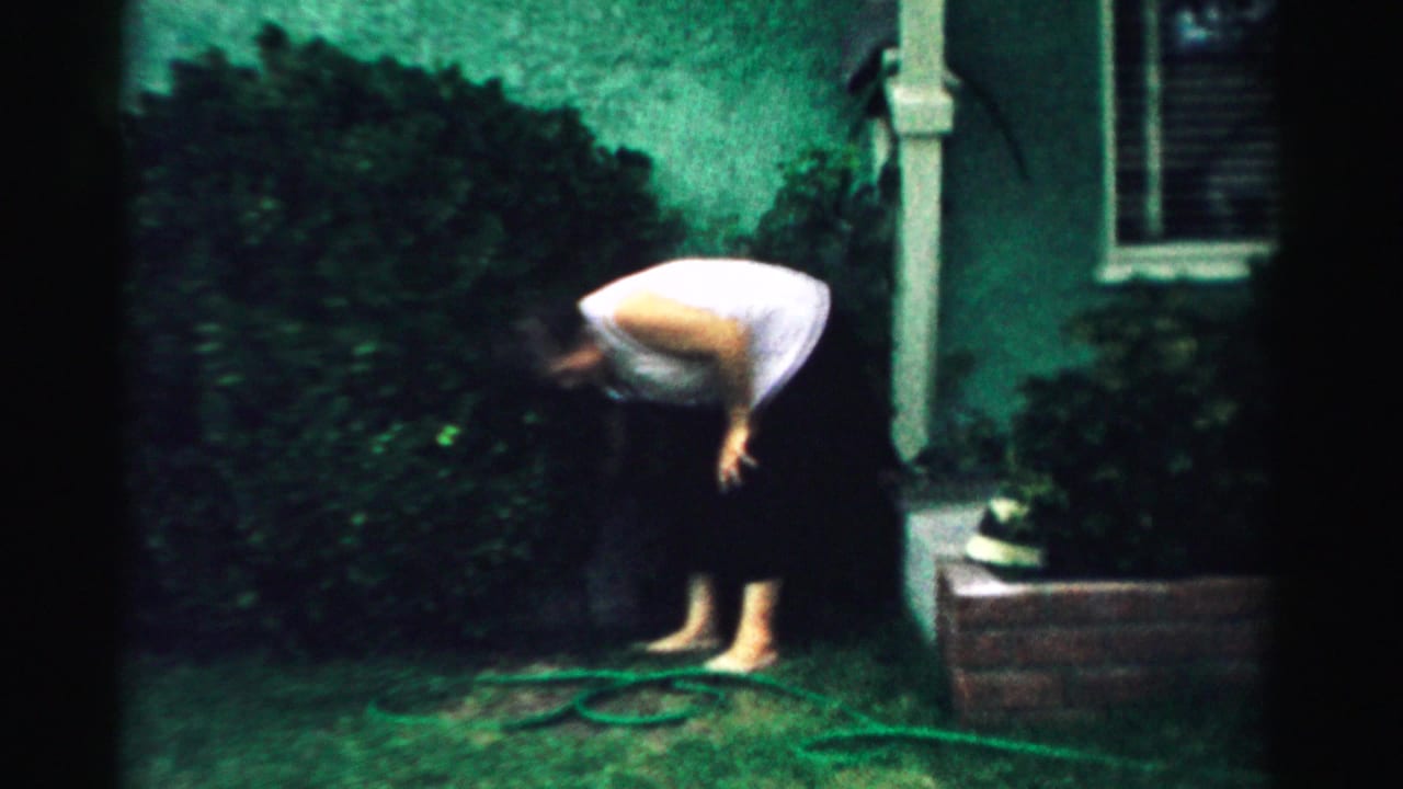 Woman bends over grass beside a coiled garden hose in a backyard.