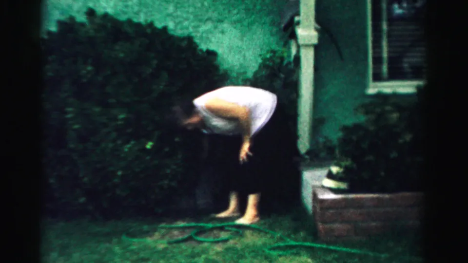 In 1957 Normal, Illinois home-movie footage, a barefoot woman in a white sleeveless top and black pants bends near a hose on green grass.
