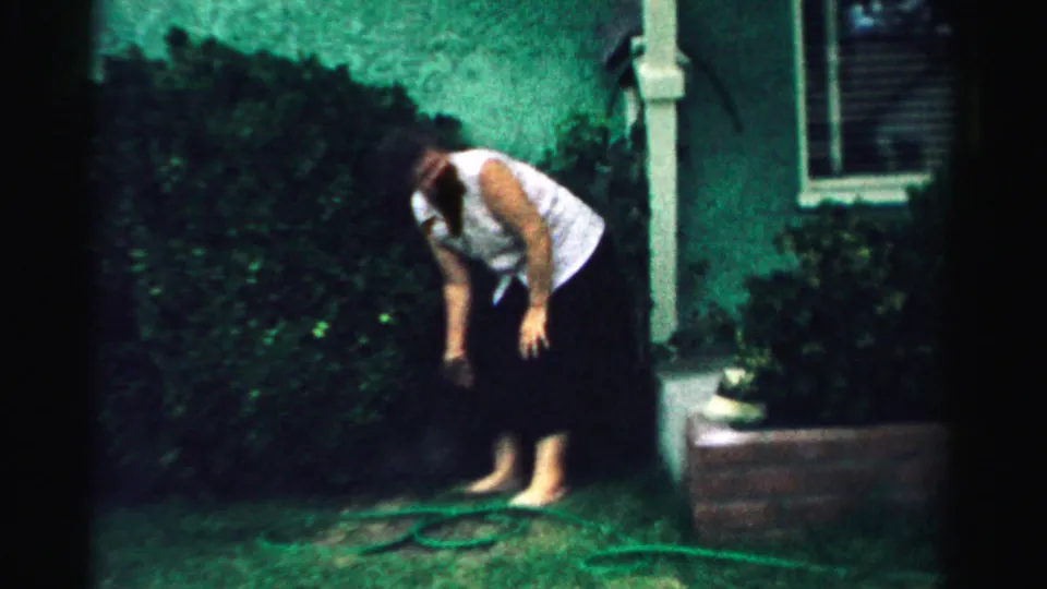 A barefoot woman bends near a garden hose in a Normal, Illinois backyard, with hedge, porch post, planter, and window visible behind her.