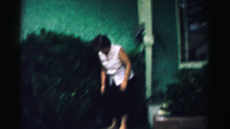 The 8mm frame shows the woman leaning forward in the backyard, with dark hedge, stucco wall, and a window partly visible in soft focus.