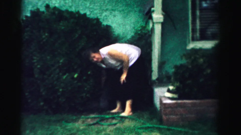 In the final frame, the barefoot woman bends over the green lawn near the hose, with a hedge, stucco house wall, and porch post in view.