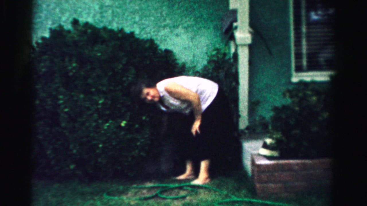 In Normal, Illinois home-movie footage from 1957, a woman in a white sleeveless top bends forward on a front lawn near a hedge.