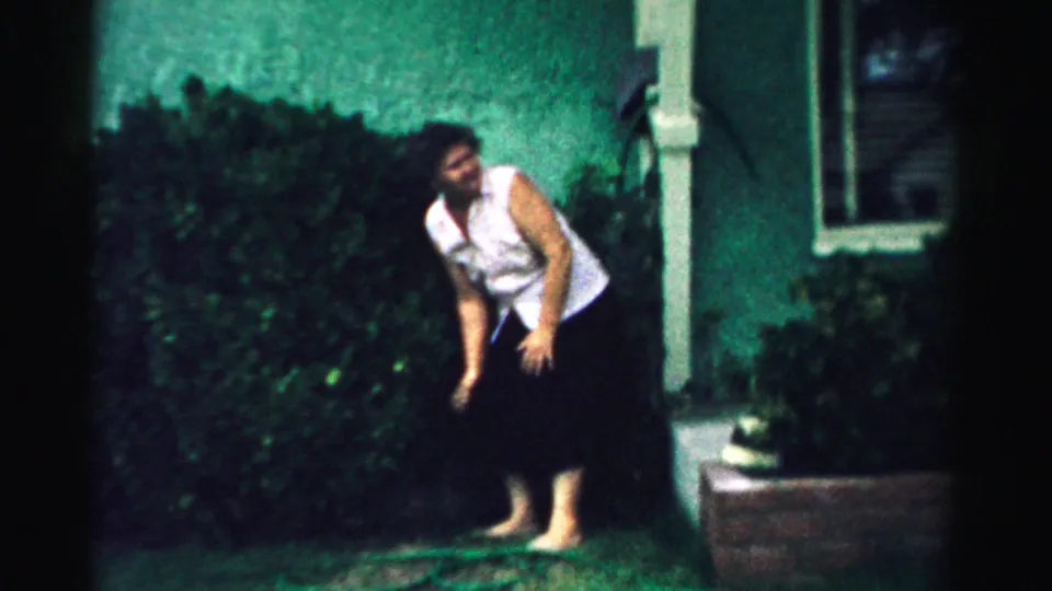 The woman pauses mid-stretch beside a green hedge and house exterior in 1957 Normal, Illinois home-movie footage.