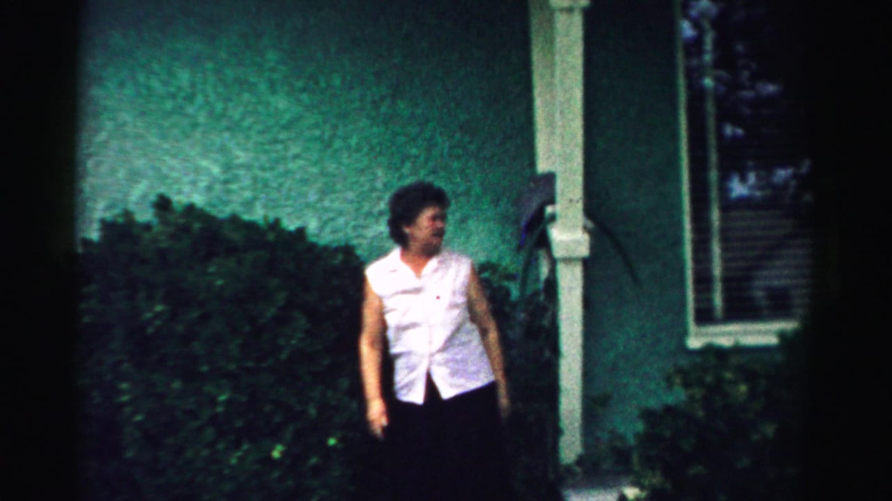 Woman in a white sleeveless blouse standing outside a green house near shrubs and a window.