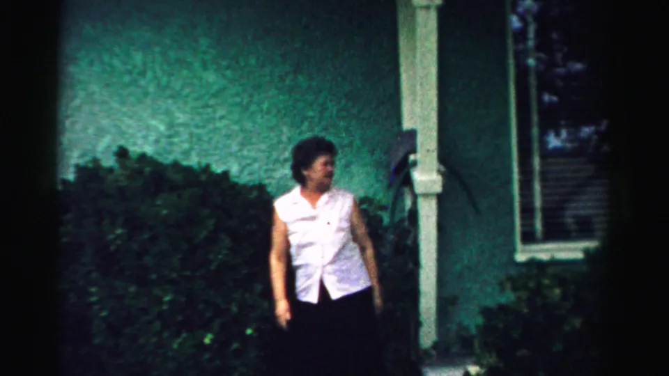 In 1957 Normal, Illinois footage, a woman stands outside a green stucco house near bushes and a window.