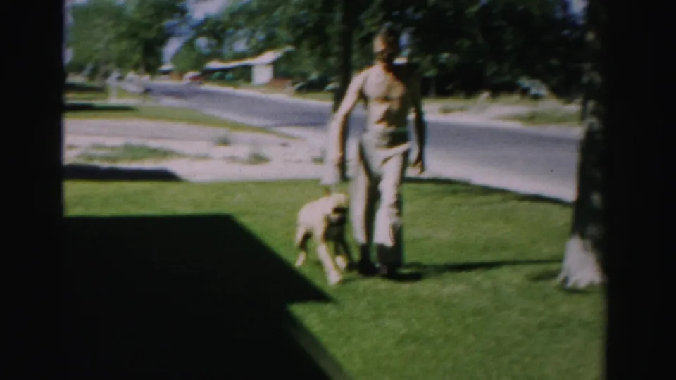 A 1957 home-movie frame shows a boy guiding a light-colored dog on a leash across a front lawn in Normal, Illinois.