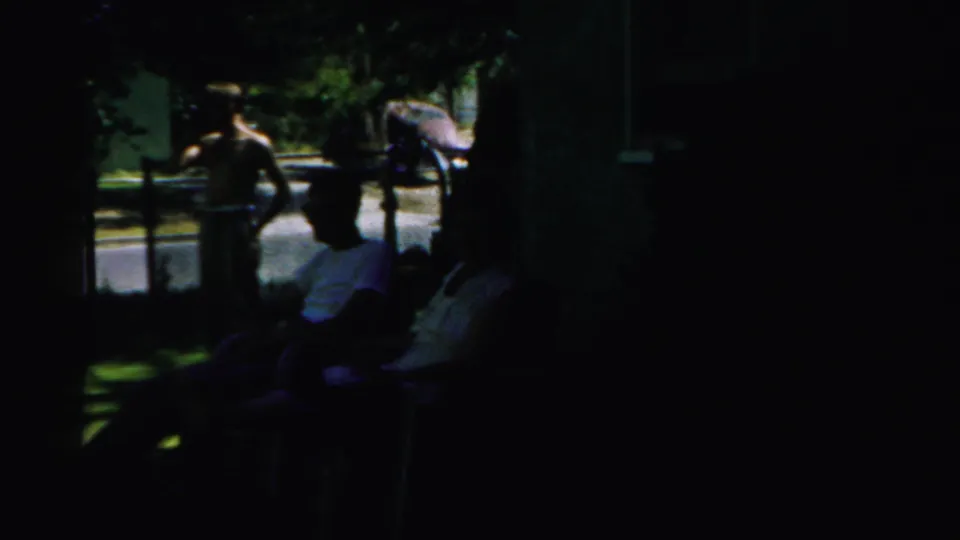 A shirtless man and leashed dog move toward neighbors seated in lawn chairs, with bright street light behind them.