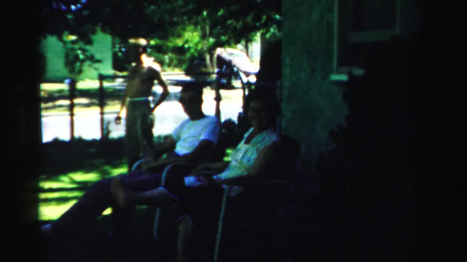 In the shaded yard, seated neighbors face a shirtless man holding a leashed dog, with trees and a bright street beyond.