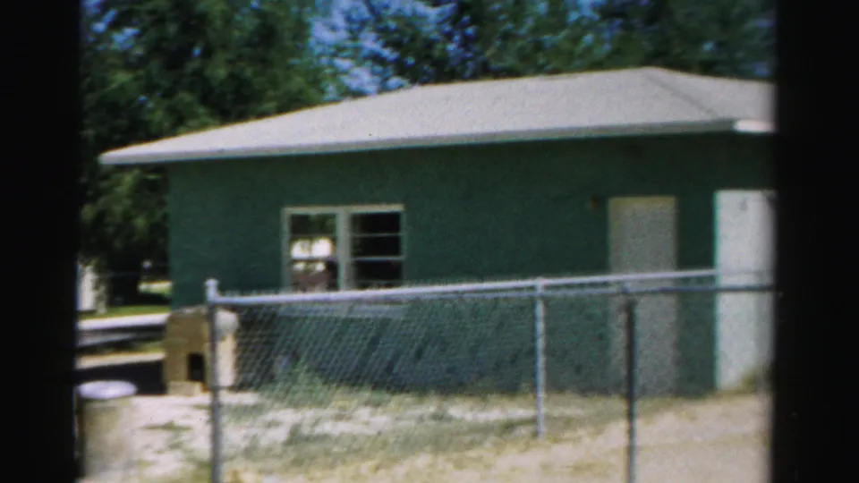 1957 Normal, Illinois exterior view of a greenish-blue building behind a chain-link fence with trees in the background.