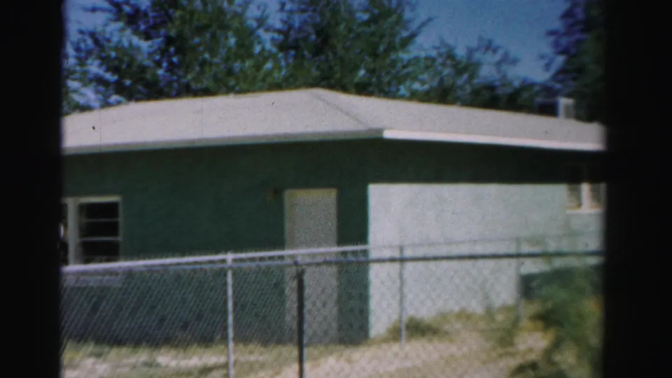 1957 8mm frame from Normal, Illinois, showing the side of a greenish-blue residential structure behind a chain-link fence.