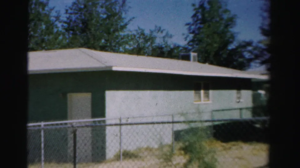 1957 Normal, Illinois home-movie frame showing a greenish-blue residential exterior with fence, yard, and leafy trees.