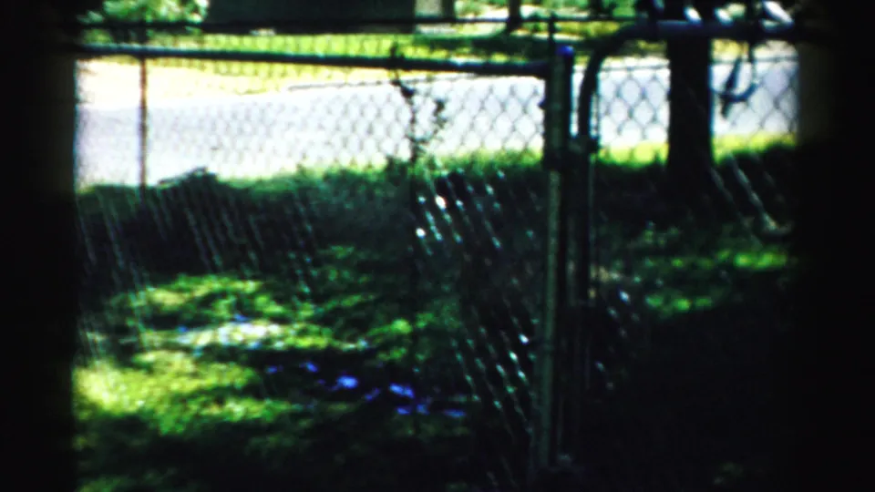 1957 Normal, Illinois home-movie frame looking through a chain-link fence toward grass and tree shade.