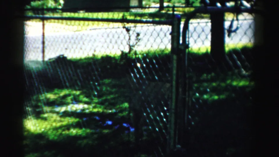 1957 Normal, Illinois frame showing a chain-link fence, shaded lawn, trees, and bright sun beyond the yard.