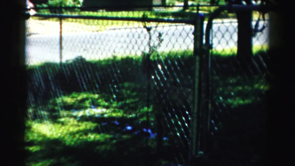 1957 Normal, Illinois home-movie frame showing a shaded yard, chain-link fence, trees, and a residential street beyond.