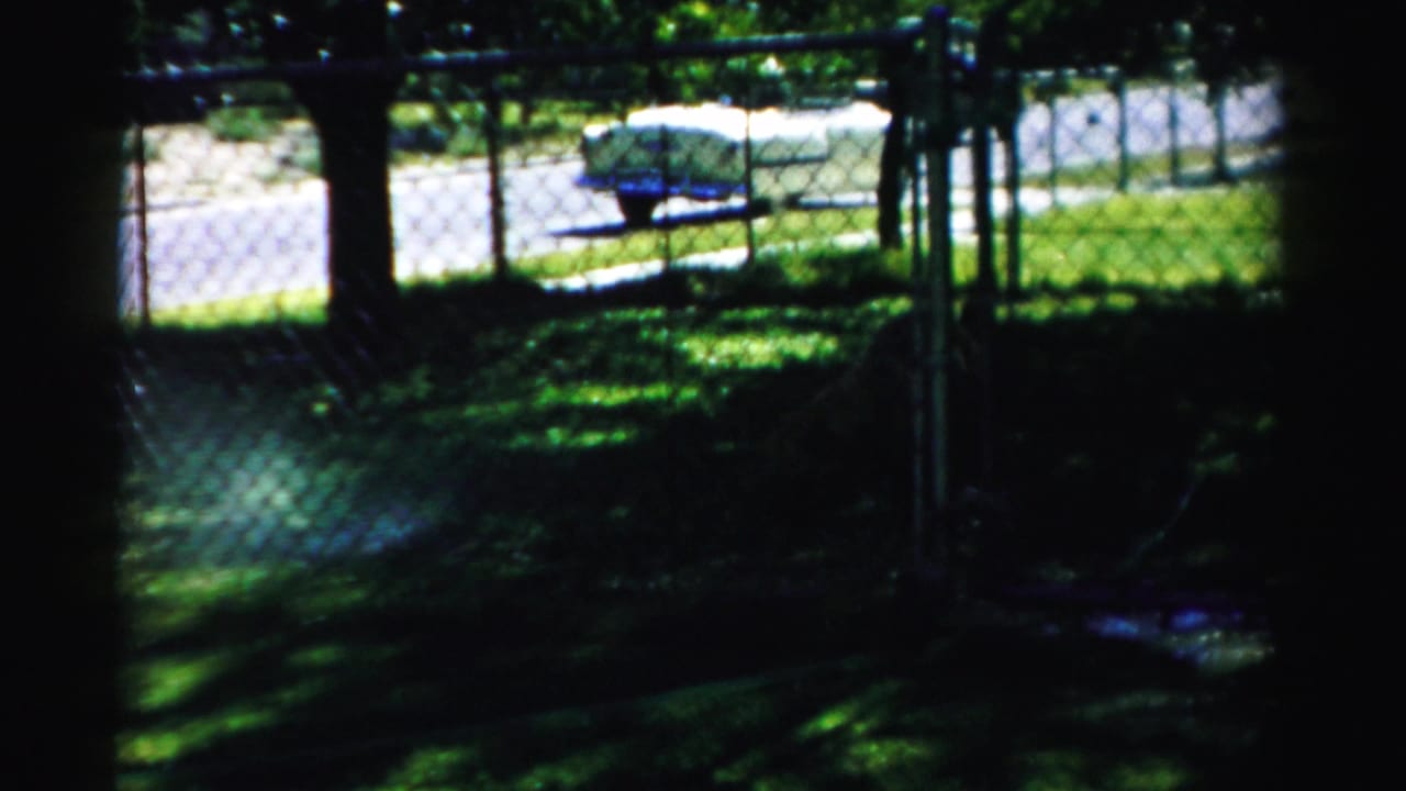 1957 Normal, Illinois home-movie frame showing a shaded yard, chain-link fence, street, and small trailer or boat-like object in bright sun.