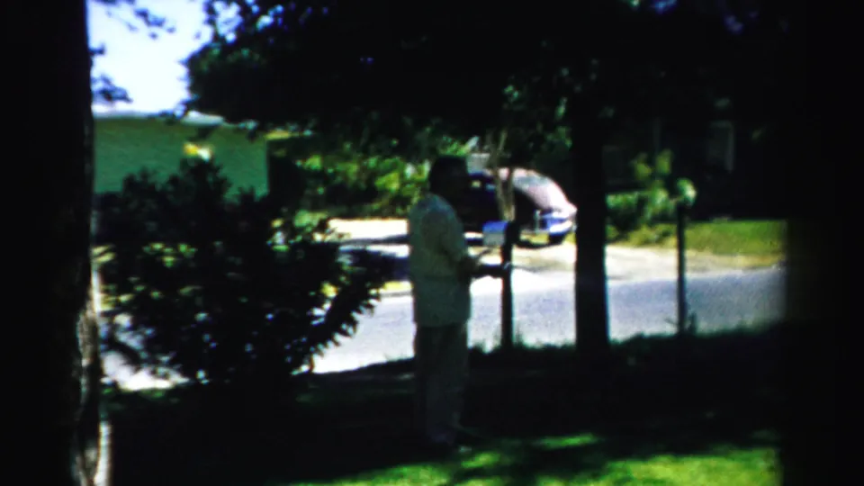1957 Normal, Illinois home-movie frame showing an adult man near a chain-link fence, with a car, street, trees, and lawn behind him.