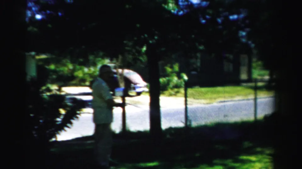 1957 Normal, Illinois frame showing an adult man watering or holding a hose in a shaded yard, with a parked car, street, and house exterior visible.