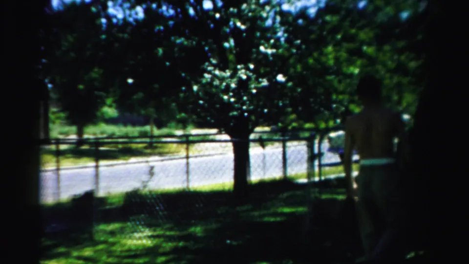 1957 Normal, Illinois yard scene with a standing man near the fence line, trees casting shade, and a street beyond.