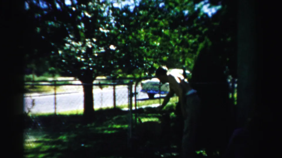 1957 Normal, Illinois home-movie frame of a man bending toward a large dog in a shaded suburban yard.