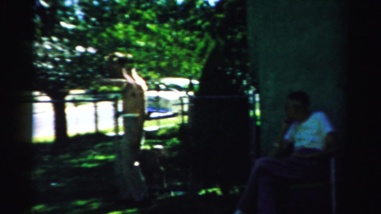 Two adults sit in shade near a house while a light-clothed figure stands in a sunny yard.