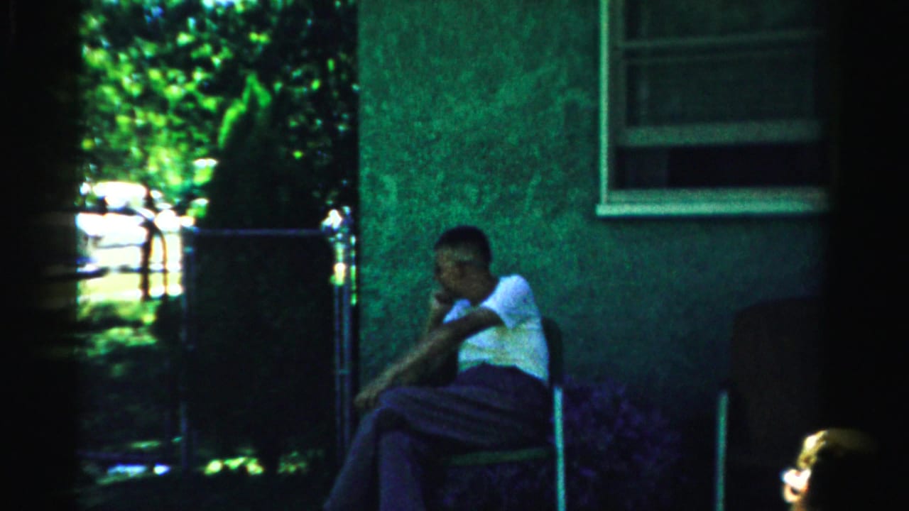 A seated man in a white shirt rests in shade beside a house wall and window.