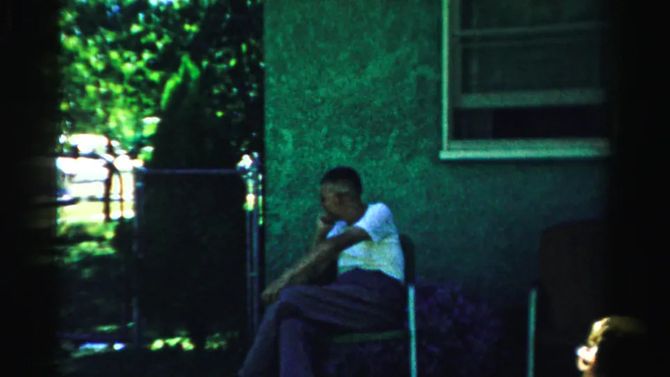 1957 Normal, Illinois frame with a seated man in shade near a residential wall, window, fence, and trees.