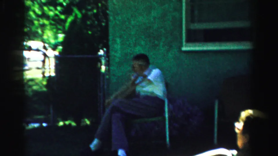 1957 Normal, Illinois home-movie frame showing a seated man in shade and a sunlit adult partially visible in the foreground.
