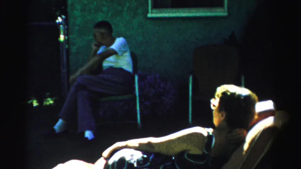 1957 Normal, Illinois frame with a shaded seated man near the house and a bright foreground adult in a chair.