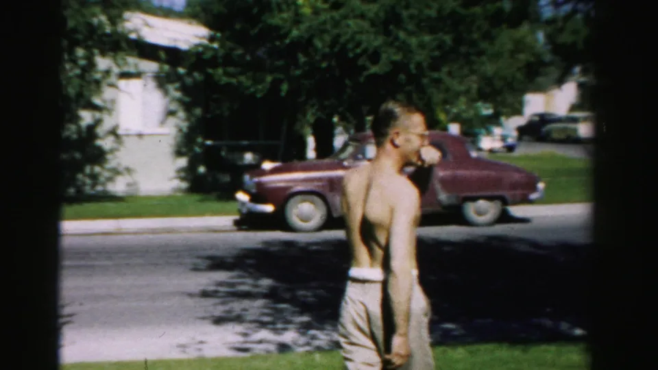 In 1957 Normal, Illinois, a shirtless man stands on grass with his back partly to camera while a maroon car sits on the residential street behind him.