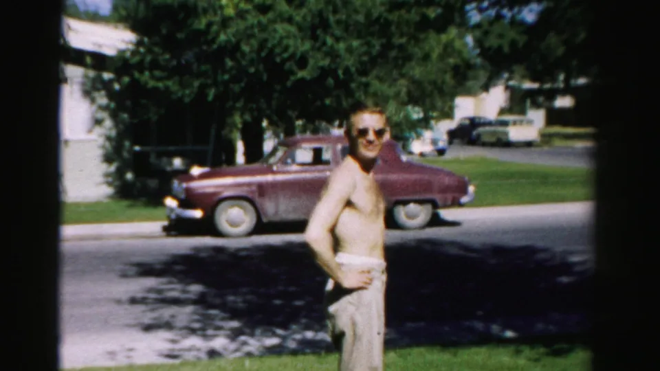 In the 1957 Normal, Illinois home movie, the shirtless man faces the camera in sunglasses beside a suburban street and maroon parked car.