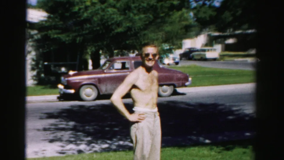 The man poses on a lawn in Normal, Illinois, wearing sunglasses and light trousers, with a maroon 1950s car parked behind him.