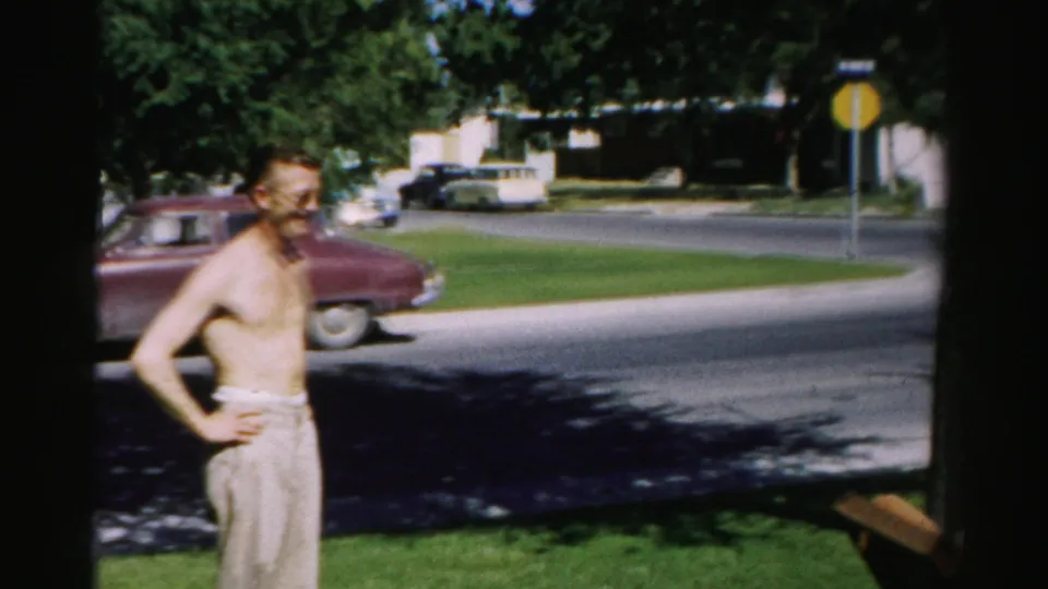 At the end of the 1957 Normal, Illinois clip, the shirtless man looks aside on the lawn with the maroon car and residential street behind him.