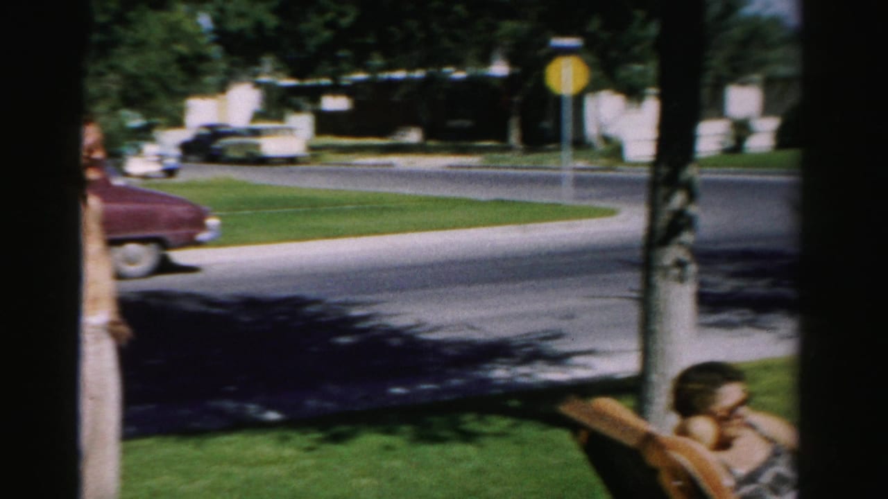 1957 Normal, Illinois home-movie frame showing a shaded front yard, standing man, reclining woman, street, red car, and yellow stop sign.