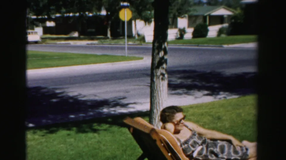 1957 Normal, Illinois frame with a woman reclining in a wooden lounge chair on a front lawn near a residential intersection.