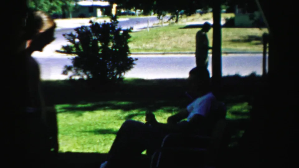 1957 Normal, Illinois home-movie frame of adults seated in shade with a bright lawn, shrub, street, and standing figure beyond.